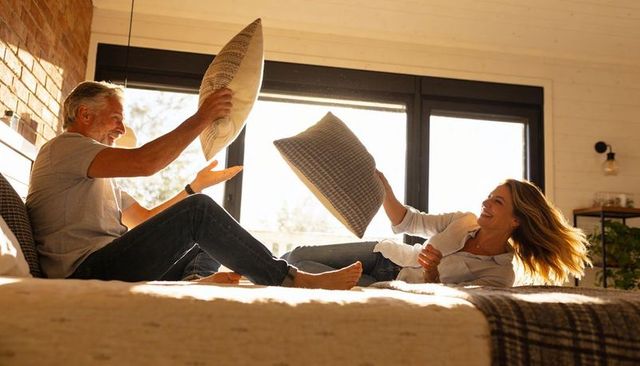 Sunlit couple pillow-fighting on cozy bed laughing and relaxing in modern bedroom