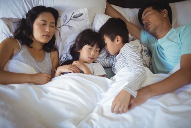 Asian family of four, including two children, sleeping together in a bed. Everyone is nestled comfortably under white blankets. Ideal for use in articles about family bonding, sleep health tips, or home living. Perfect for illustrating concepts of relaxation, comfort, and familial love.