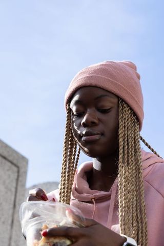 African american teen wearing pink beanie holding clear food container in urban plaza