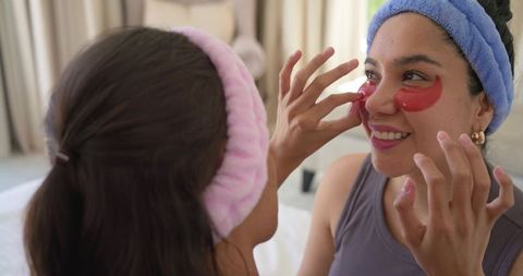 Friends applying red under-eye gel patches while smiling on bed at home during skincare routine