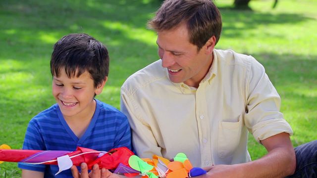 Father and son enjoying each other's company while looking at a colorful kite. Great for themes related to family bonding, outdoor activities, leisure time, and parent-child relationships. Perfect for advertisements, social media posts, and websites promoting family-oriented activities.