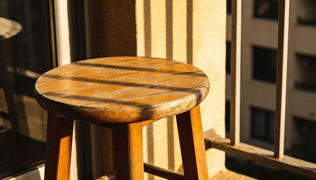 Sunlit weathered wooden stool on urban balcony casting diagonal shadow strips at golden hour