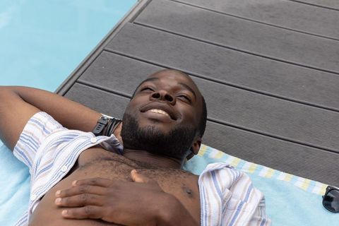 African american man reclining poolside on blue towel wearing striped open shirt and watch