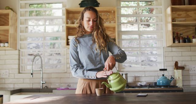Young woman preparing tea with a green teapot in a modern kitchen with wooden shelves and ample sunlight. Suitable for themes around domestic life, morning routines, relaxation, and home activities. Useful for lifestyle blogs, cooking websites, and interior design showcases.