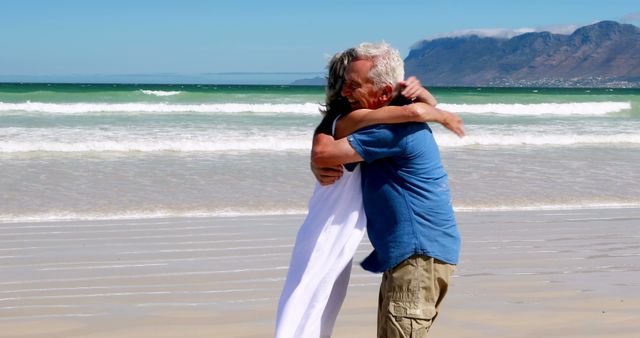 Senior couple hugging joyfully on a sunny beach with ocean waves and distant mountains in background. Great for use in contexts of retirement lifestyle, love stories, travel promotions, or advertisements emphasizing happiness in older age.