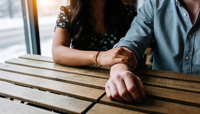 Couple holding hands on wooden table in sunlit cafe, intimate cozy romance