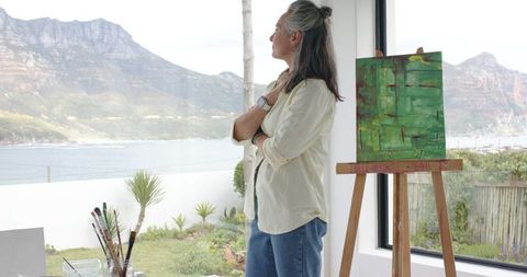 Mature artist gazing over coastal bay with easel and abstract green canvas