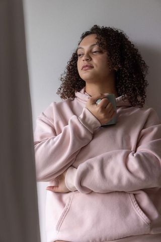 Contemplative african american woman holding mug in pink hoodie by window