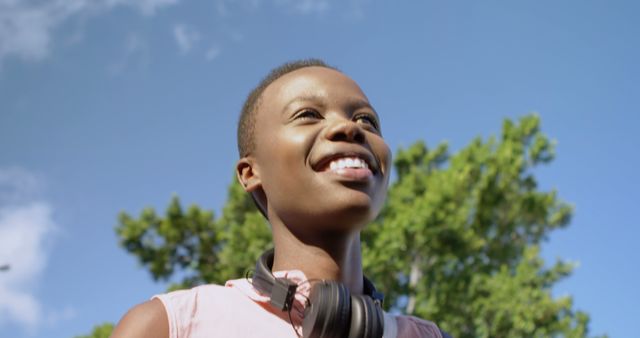 This photo of a smiling young woman wearing headphones against a backdrop of clear blue sky and lush green trees can be used in advertising campaigns, social media posts focused on happiness and wellness, websites promoting outdoor activities, and educational content about the positive effects of nature and music.