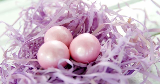 Three pastel pink eggs rest in a bed of shredded purple paper, symbolizing Easter or springtime. The soft colors and seasonal theme make it ideal for festive decoration or holiday-related marketing.