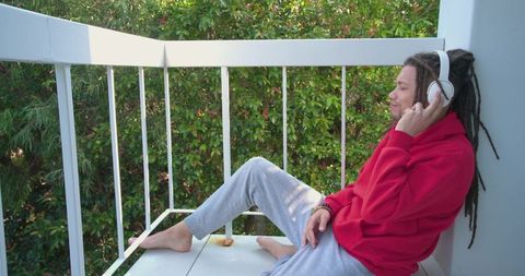 Young man with dreadlocks listening to music on balcony in red hoodie and white headphones