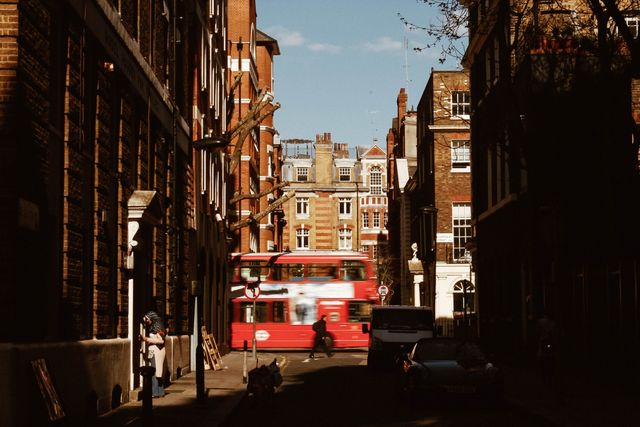 Photo capturing a busy urban scene in a historic alleyway in London, featuring a traditional red double-decker bus in motion and a blend of old and modern architecture. Suitable for travel blogs, websites promoting tourism, educational articles on British culture and urban development, or marketing materials for luxury urban living.