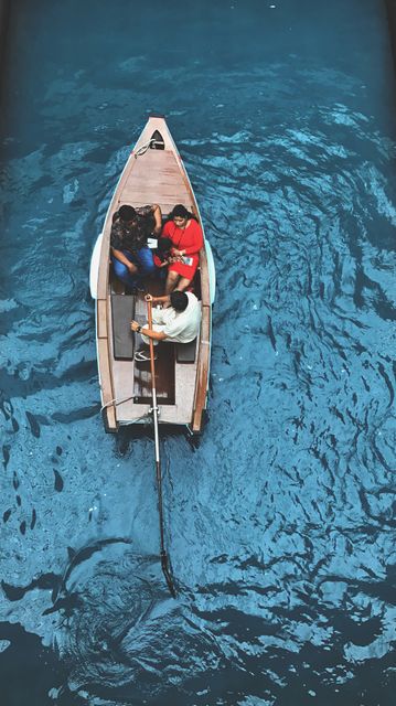 Top view of a group of people enjoying a boating experience in a wooden rowboat. They are navigating through vibrant blue water, showcasing a leisurely and adventurous water activity. This scene can be used to depict themes of travel, exploration, and outdoor fun, perfect for tourism promotions, outdoor activity campaigns, and leisure lifestyle imagery.