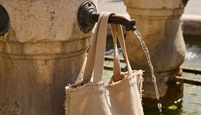 Hanging beige woven tote bag on fountain spout with flowing water and weathered stone