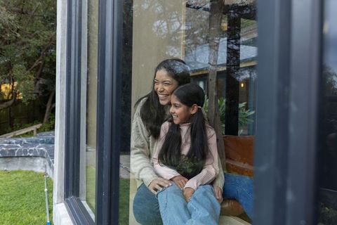 Indian mother and daughter sitting by window smiling and looking at garden