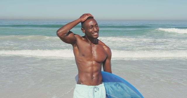 Smiling african american man carrying surfboard walking on sunny beach by the sea. Summer, hobbies, surfing and vacations.