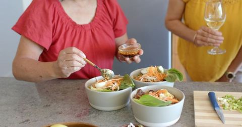 Diverse senior women preparing healthy salad bowls and enjoying wine in home kitchen