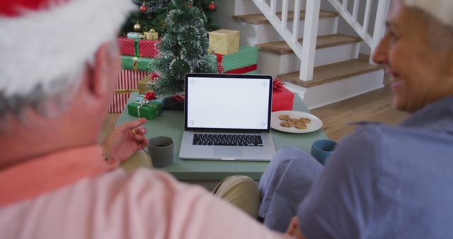 Senior couple engaging in a video call while sitting in front of a laptop. Background features Christmas tree with decorations, wrapped presents, and plate of cookies on the table. Suitable for themes such as holiday celebrations, family connections, and festive digital communication.