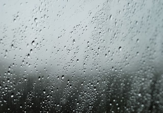 This image captures raindrops clinging to a window glass, blurring the view of an overcast, gloomy outdoor setting. The droplets create a pattern and reflect light subtly. Perfect for themes on weather, mood, serenity, reflection, and nature visuals on websites, blogs, or social media posts conveying tranquility, melancholy, or stormy weather vibes.