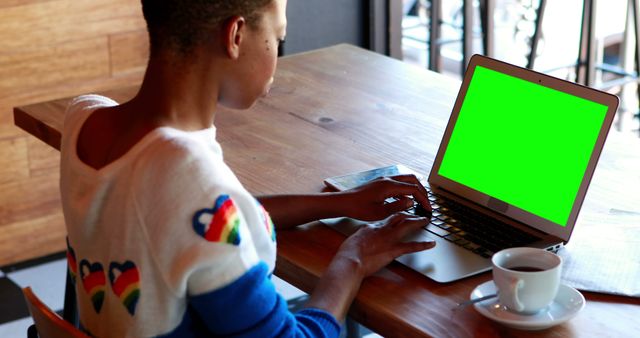 Individual working on a laptop with a green screen in coffee shop. Suitable for illustrating remote work, freelance lifestyle, technology use in public places, and similar modern-day scenarios.