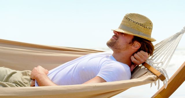 A man wearing a white t-shirt and a straw hat is relaxing on a hammock at the beach. The serene coastal setting and the laid-back posture of the man suggest a moment of peaceful leisure, ideal for promoting travel, summer vacation destinations, relaxation techniques, or lifestyle blogs focused on mindful living.