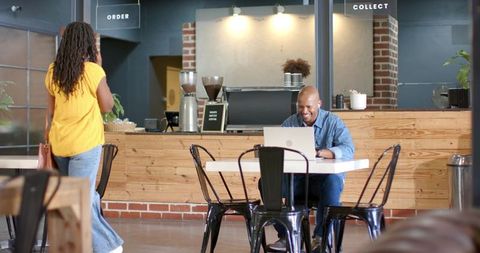 African american man working on laptop in modern industrial coffee shop while woman orders