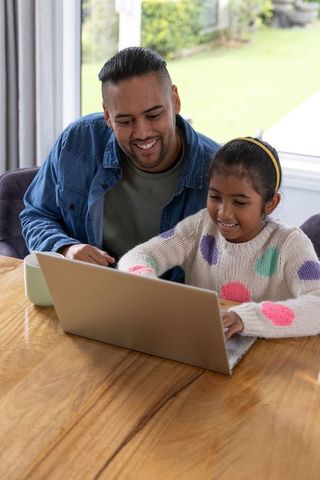 Asian father guiding daughter using laptop for online learning at home kitchen table