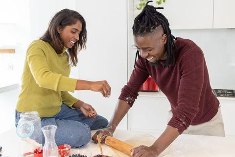 Diverse couple baking together on kitchen island, woman sprinkling flour, man rolling dough
