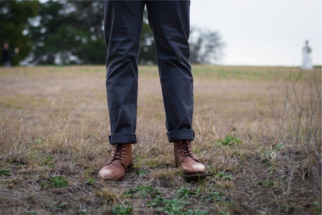 Close-up of a man wearing stylish brown leather boots and rolled up gray pants standing in a dry, grassy field. This image can be used for content related to men's fashion, outdoor activities, fall styles, and casual footwear.