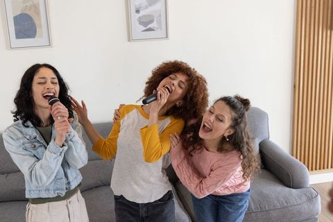 Diverse friends singing karaoke at home, joyful trio bonding in cozy modern living room
