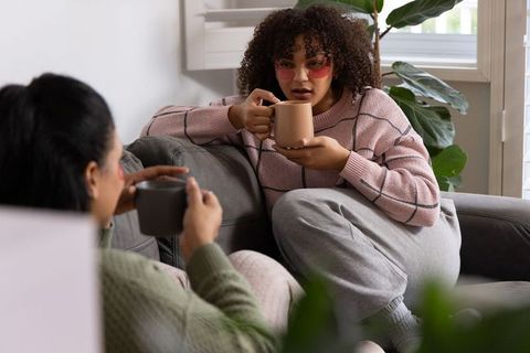 Diverse female friends chatting on cozy sofa sipping coffee and wearing under-eye patches
