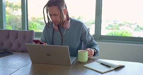 Remote worker concentrating at laptop with smartphone and coffee, headphones around neck