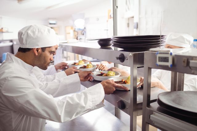 Chefs in a commercial kitchen passing ready-to-serve dishes to a waiter at the order station. Ideal for use in articles or advertisements related to the restaurant industry, culinary arts, teamwork in professional kitchens, or hospitality services.