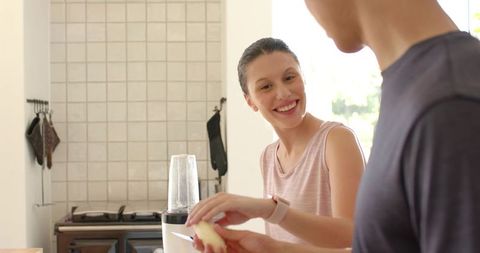 Smiling woman watching partner slicing apple beside blender on sunlit modern kitchen counter
