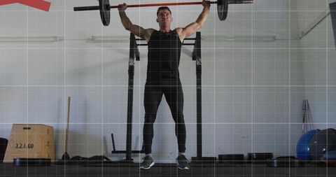 Male weightlifter pressing loaded barbell overhead in garage gym wearing black gear