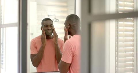 African American man smiling at mirror applying skincare in bright modern bathroom