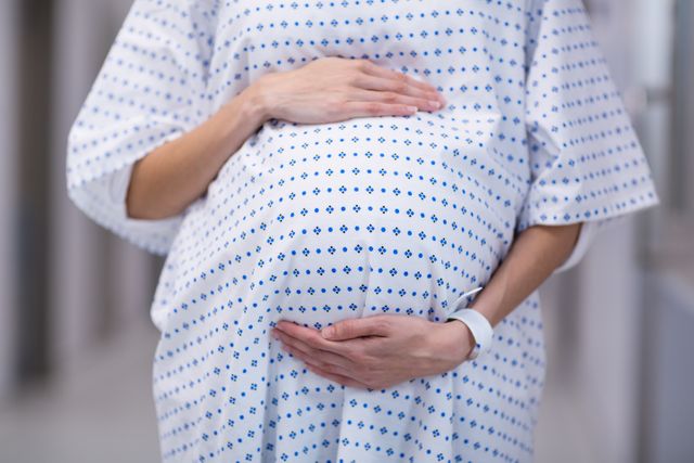 This image shows a pregnant woman standing in a hospital corridor, gently holding her belly. She is wearing a medical gown, indicating she is likely in a healthcare setting for a prenatal checkup or preparing for delivery. This image can be used for articles, blogs, or advertisements related to maternity care, prenatal health, pregnancy, and hospital services.