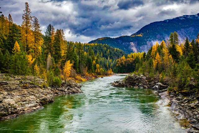 Tranquil river meandering through a lush forest with vibrant autumn foliage, set against a breathtaking mountain backdrop under a dramatic cloudy sky. Ideal for use in travel brochures, nature-themed projects, environmental campaigns, and as background imagery for websites promoting outdoor activities or serene landscapes.