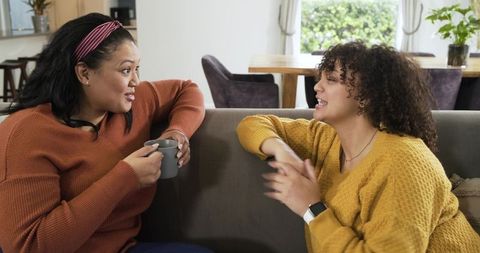 Two friends having cozy conversation on gray sofa wearing knit sweaters holding mug