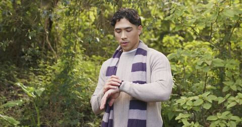 Asian man checking smartwatch while standing in sunlit forest wearing striped scarf and sweater