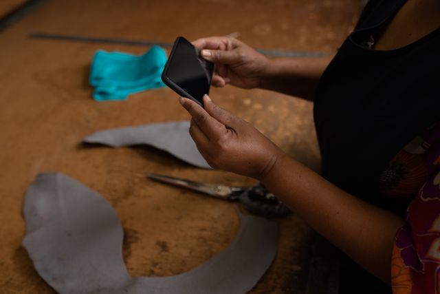 Side view close up of hands of a biracial woman using a smartphone at a hat factory. In the background bales of material can be seen in storage.