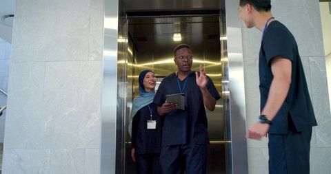 Diverse healthcare team exiting elevator holding tablet and greeting colleague in hospital