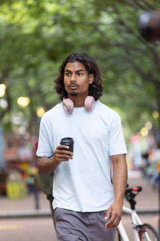 Indian man walking with coffee and pink headphones on tree-lined urban street
