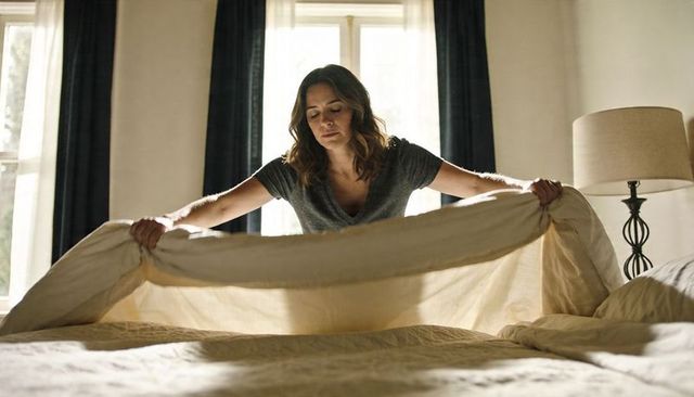 Woman making bed in sunlit bedroom smoothing beige duvet over mattress with bedside lamp