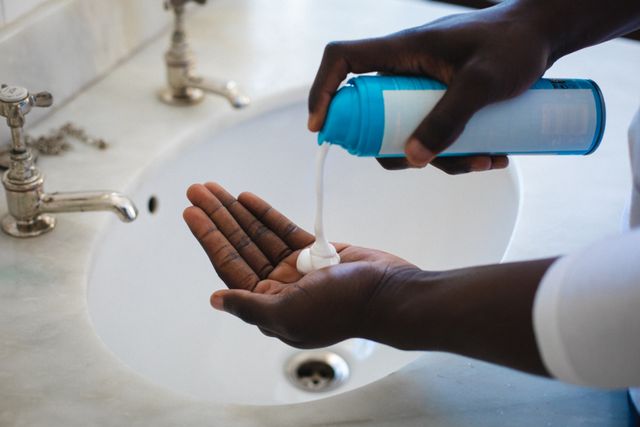 This image shows an African American man applying shaving foam over a bathroom sink. It is ideal for use in advertisements or articles related to personal grooming, skincare routines, hygiene products, and lifestyle blogs. The focus on hands and the shaving foam makes it suitable for illustrating tutorials or product demonstrations.