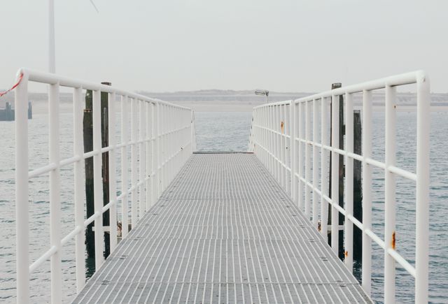 View of an empty metal walkway extending over calm water leading to the horizon on an overcast day. Ideal for use in travel blogs, websites focusing on nature and tranquility, or materials highlighting paths and journeys.