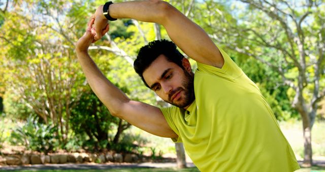 Man in yellow shirt stretching in park surrounded by trees and greenery. Use for fitness, healthy lifestyle, outdoor exercise, workout routines, wellness, and nature-related content.