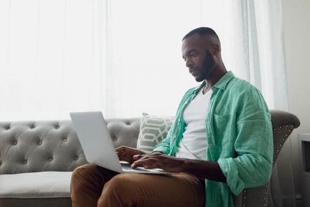 African-American man sitting on a grey couch using a laptop. Ideal for illustrating remote work, home office setups, casual work environments, and technology use in everyday life.