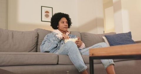 African american man lounging on sofa at home eating popcorn watching tv in denim jacket