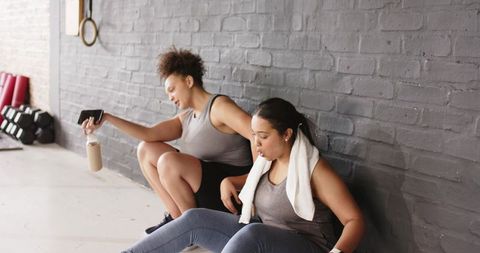 Diverse women resting and checking smartphone after intense gym workout, cooling down